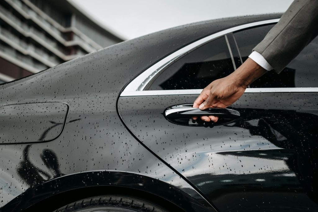 A close-up of a hand opening a black car door covered in raindrops, showcasing elegance and weather detail.